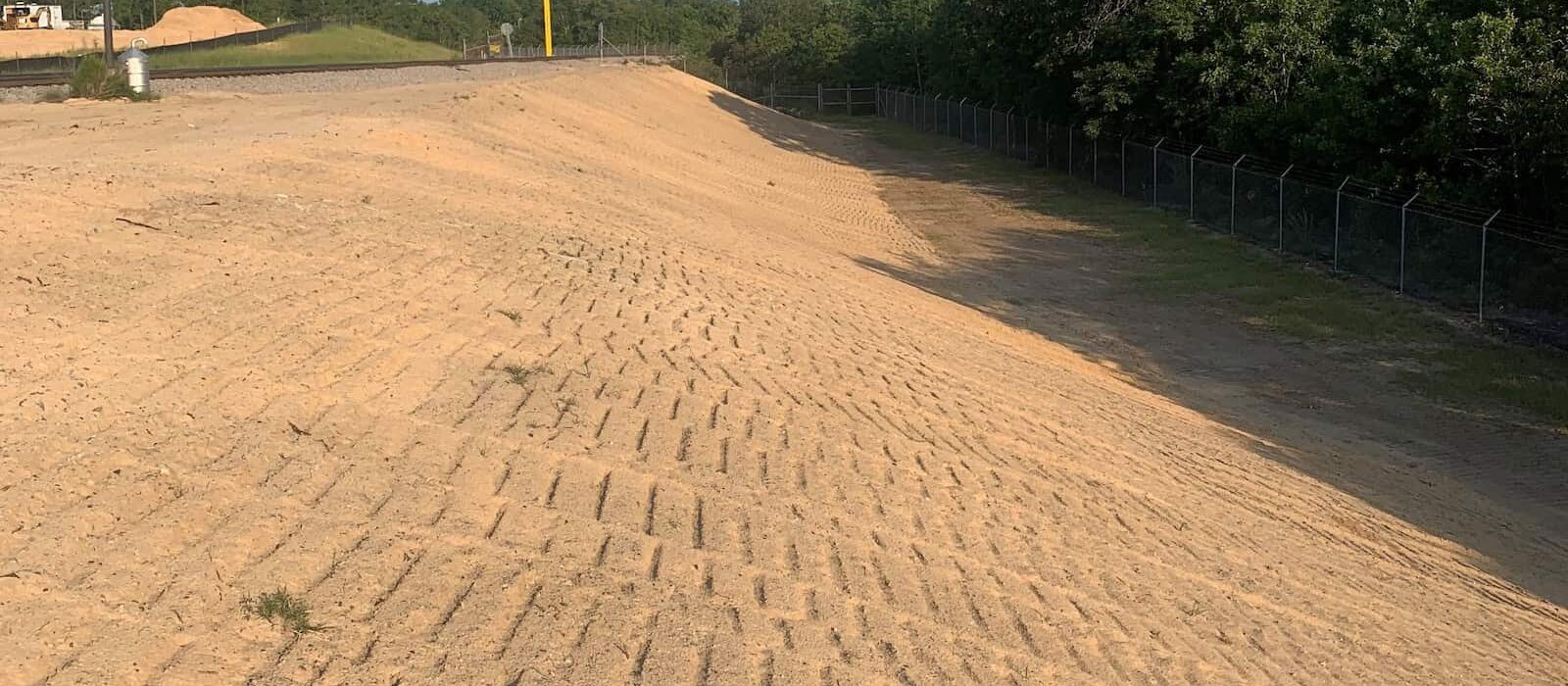 Graded sandy slope with track marks near an industrial facility, bordered by trees and fencing.