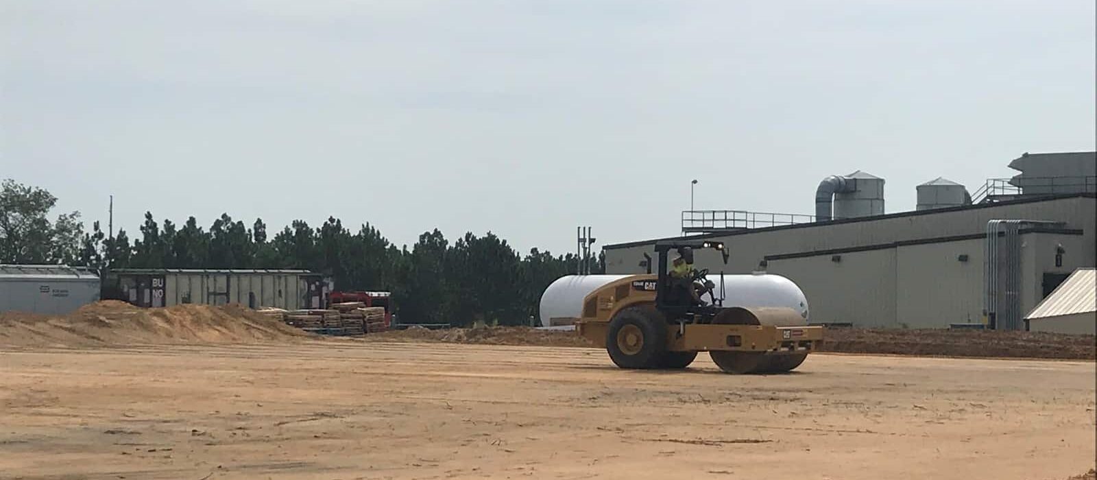 Worker operating a soil compactor on a cleared construction pad next to a large manufacturing building.