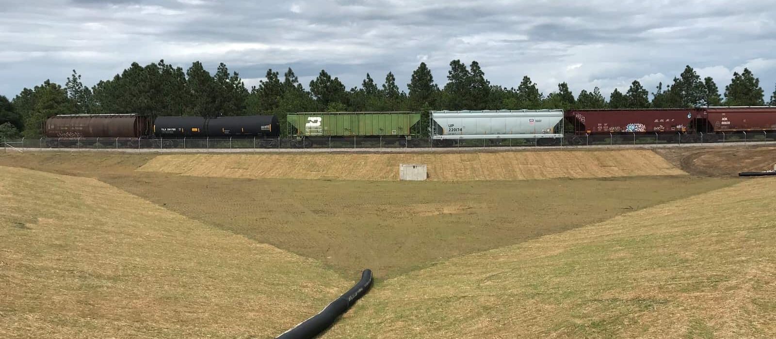 Grassed stormwater detention basin with a drain pipe, adjacent to a rail line with freight train cars.