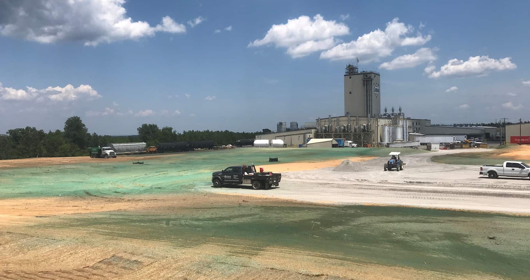 Large hydroseeded field next to an industrial plant with work vehicles and storage tanks on-site.