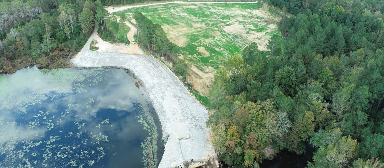 Wide-angle aerial image showing lake, dam, spillway, riprap reinforcement, and large re-vegetated area adjacent to the forest.