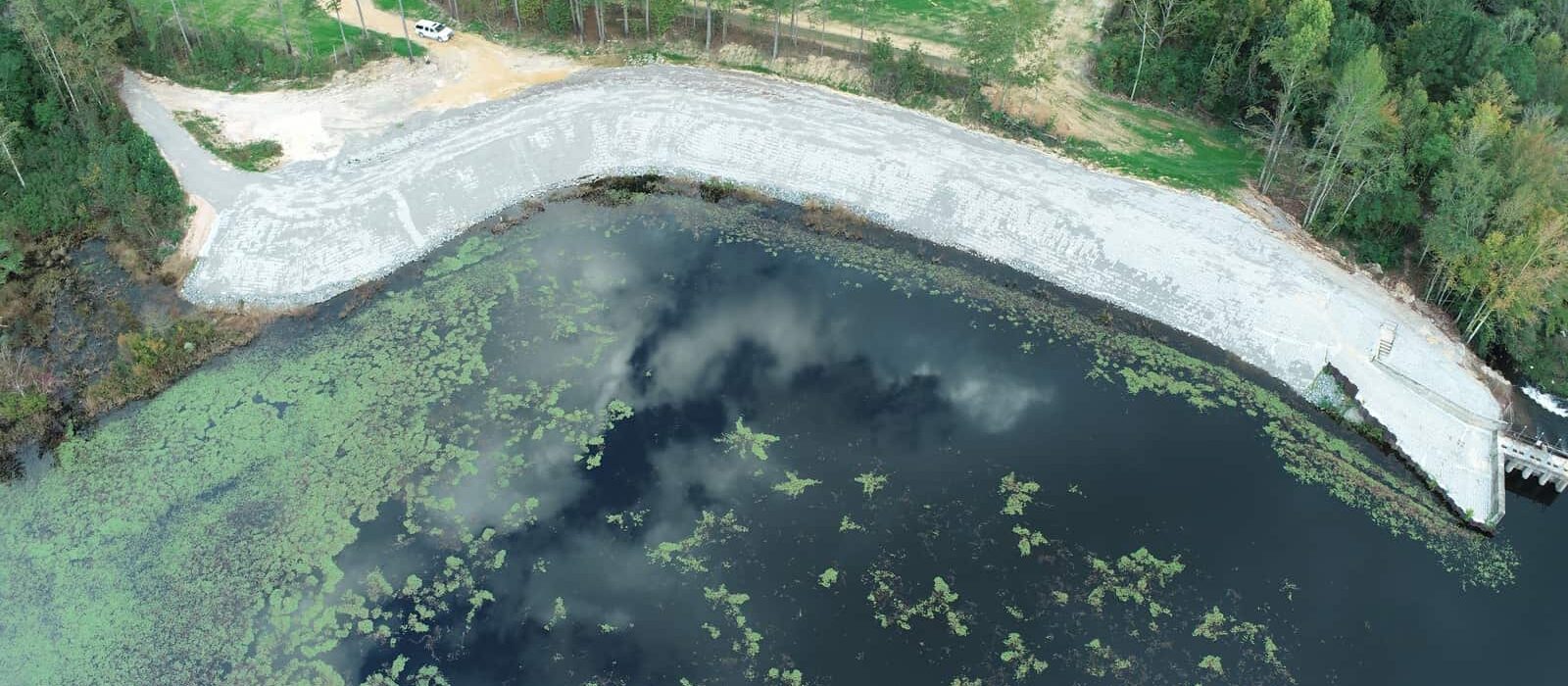 Overhead view of a lakebank lined with riprap and a visible dam, adjacent to a newly seeded field and wooded area.