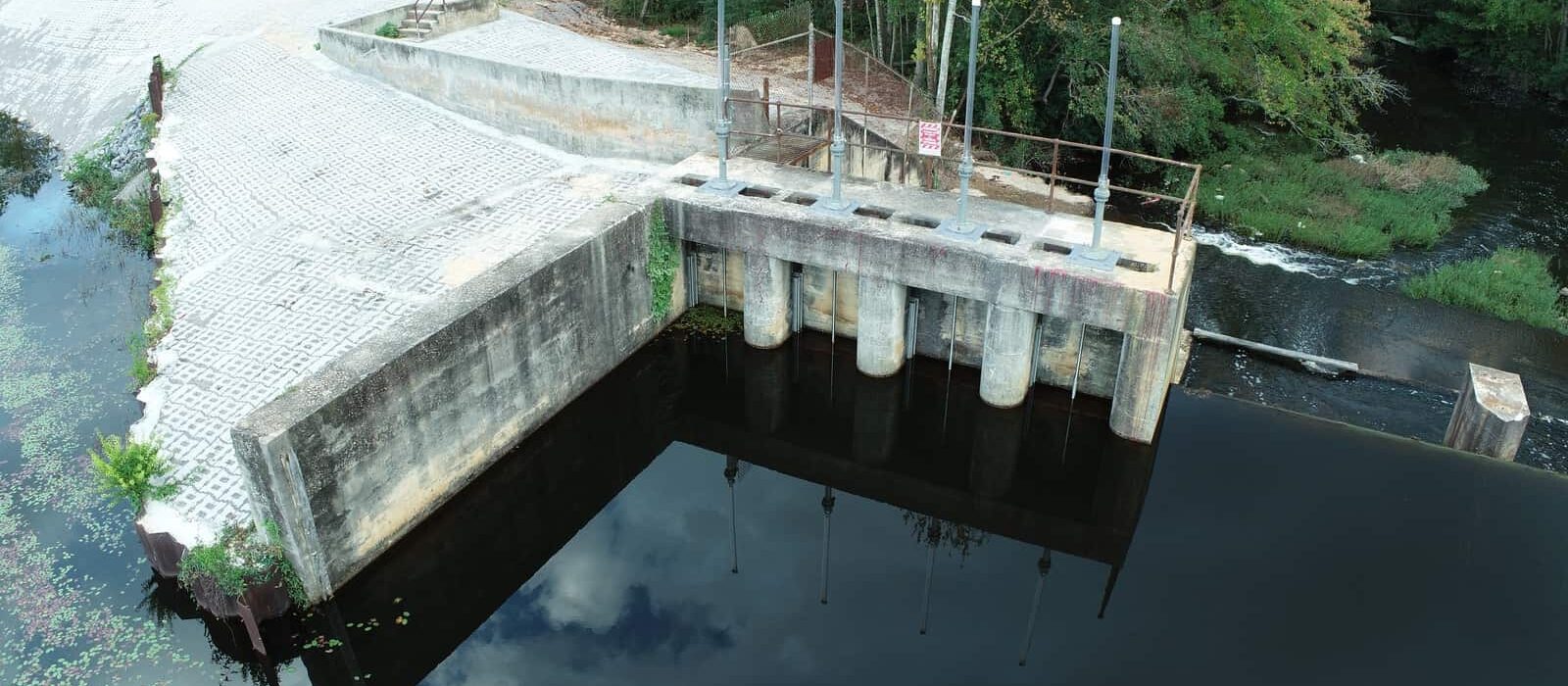 Close-up of concrete dam with metal gate controls and reinforced embankment, showing spillway and surrounding forest.