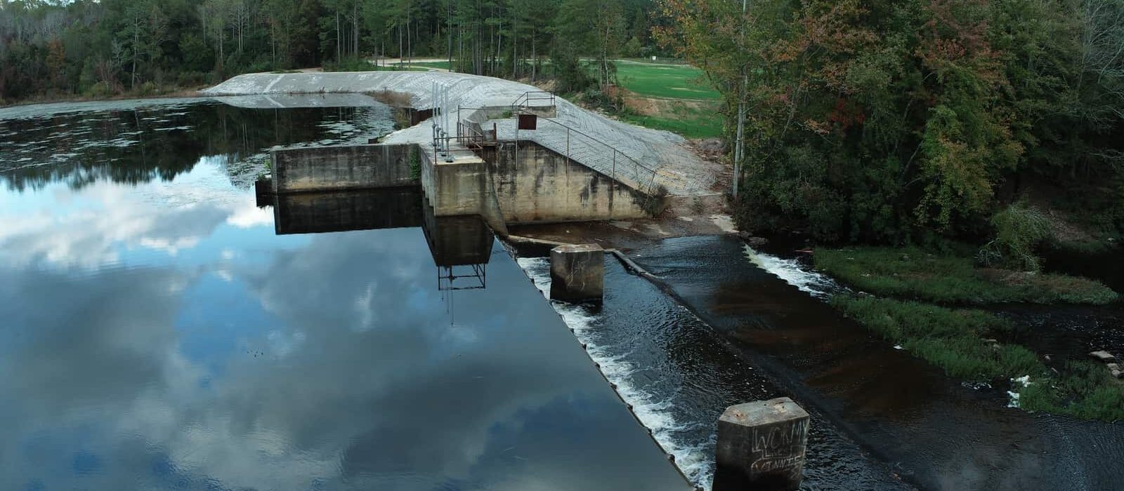 Side view of dam and spillway where water flows over into a creek, surrounded by dense trees and reflections on the water.
