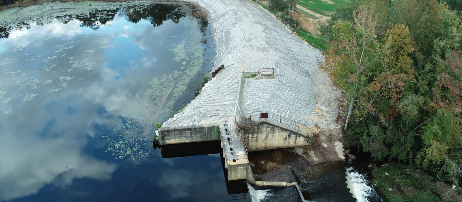 Elevated view of dam outlet and spillway with water release visible and embankment protected by grid-patterned concrete blocks.