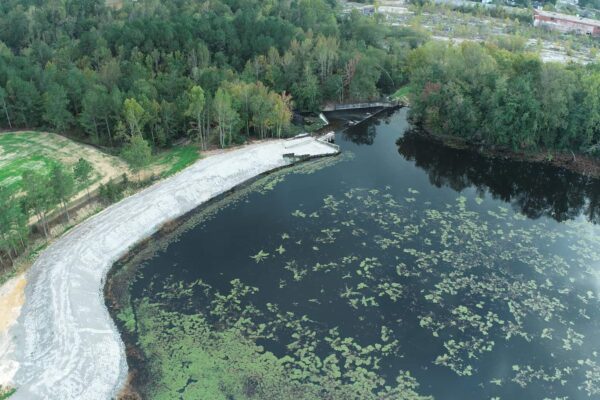 Aerial view of a forested lake with visible dam structure and riprap slope reinforcement along the shoreline.