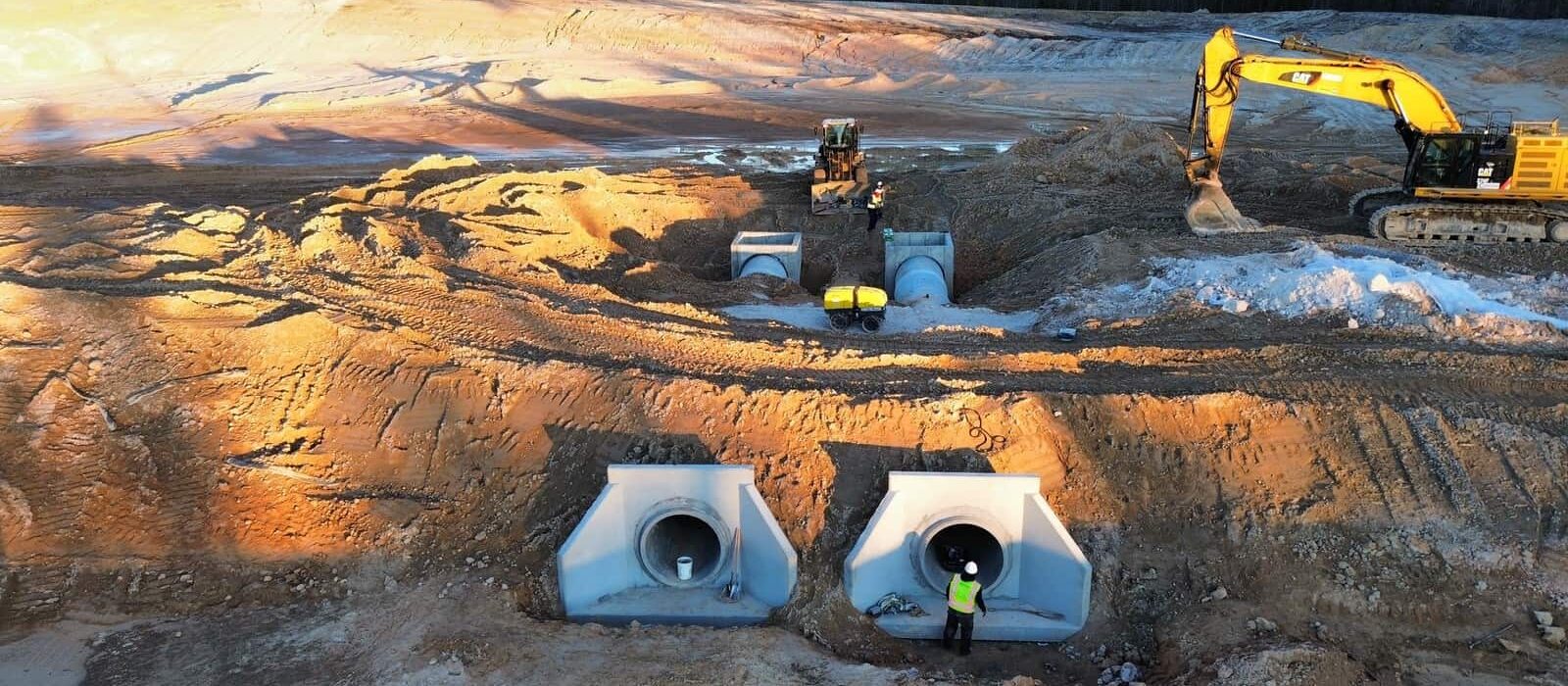 Excavator and crew installing large concrete drainage pipes at a construction site with early morning sunlight.