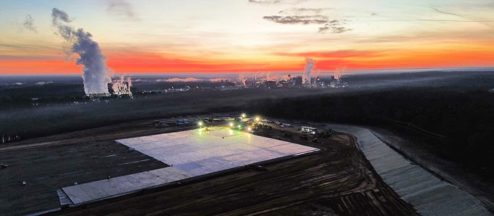Aerial view of an industrial construction site at dawn with site lighting and factory smoke stacks in the background.