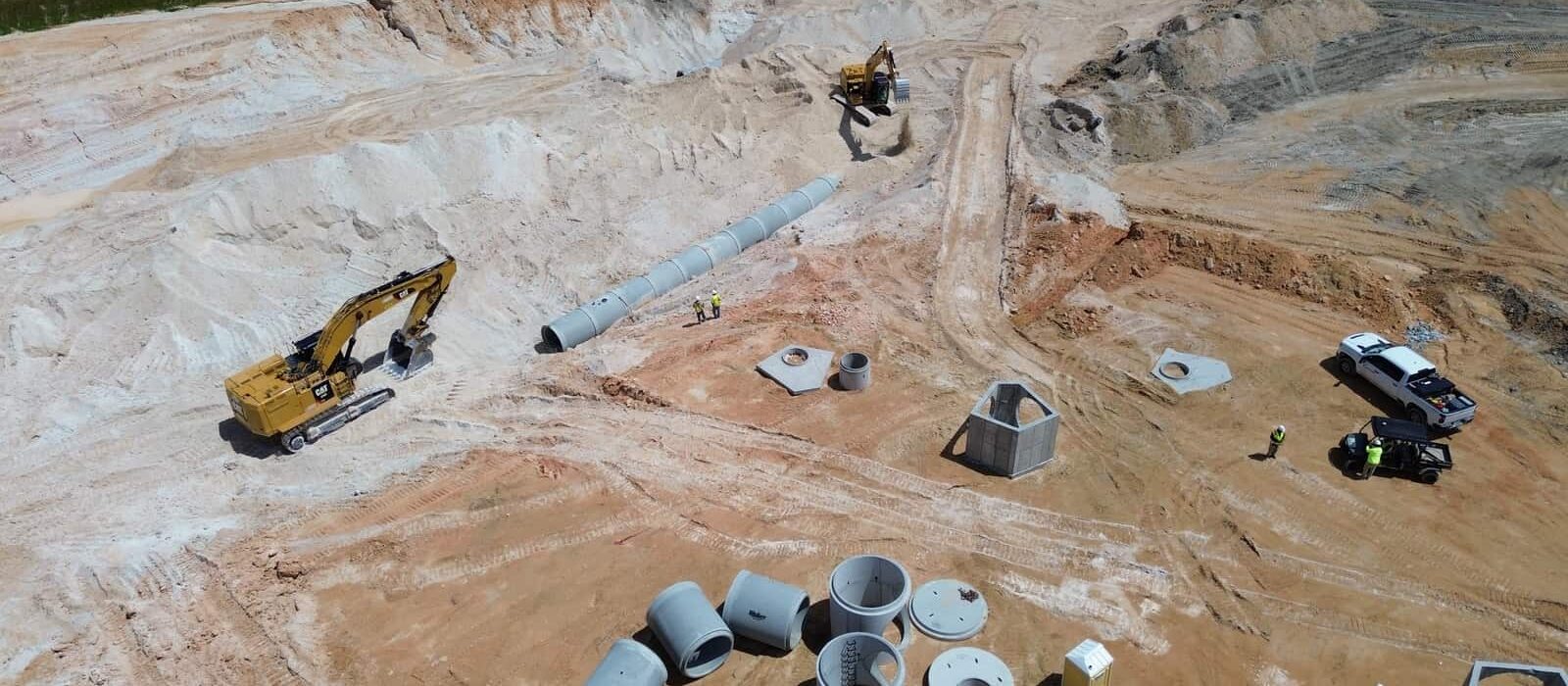Excavators and workers installing large concrete drainage pipes and structures in a deep excavation at a construction site.