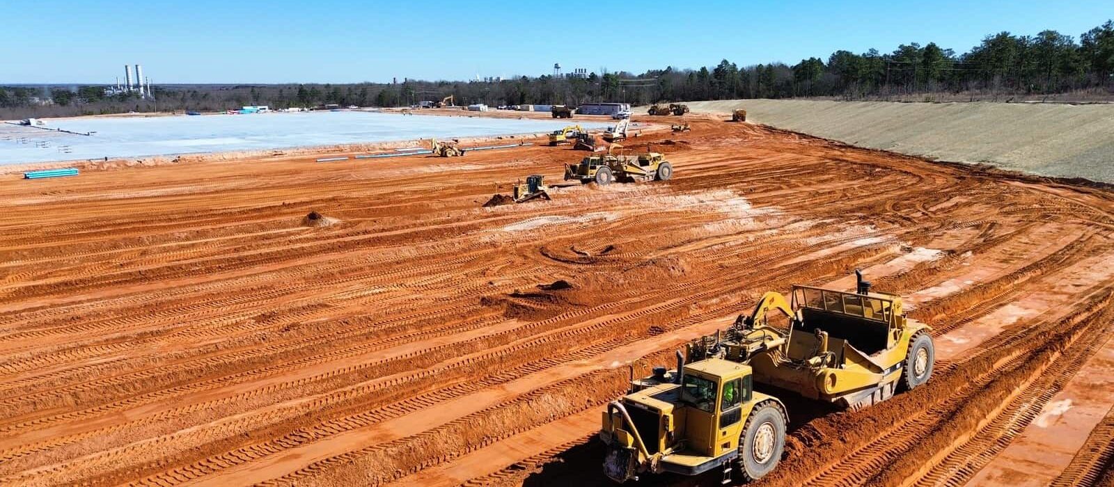Scraper machines grading red clay at a large construction site with concrete slab work in the background.