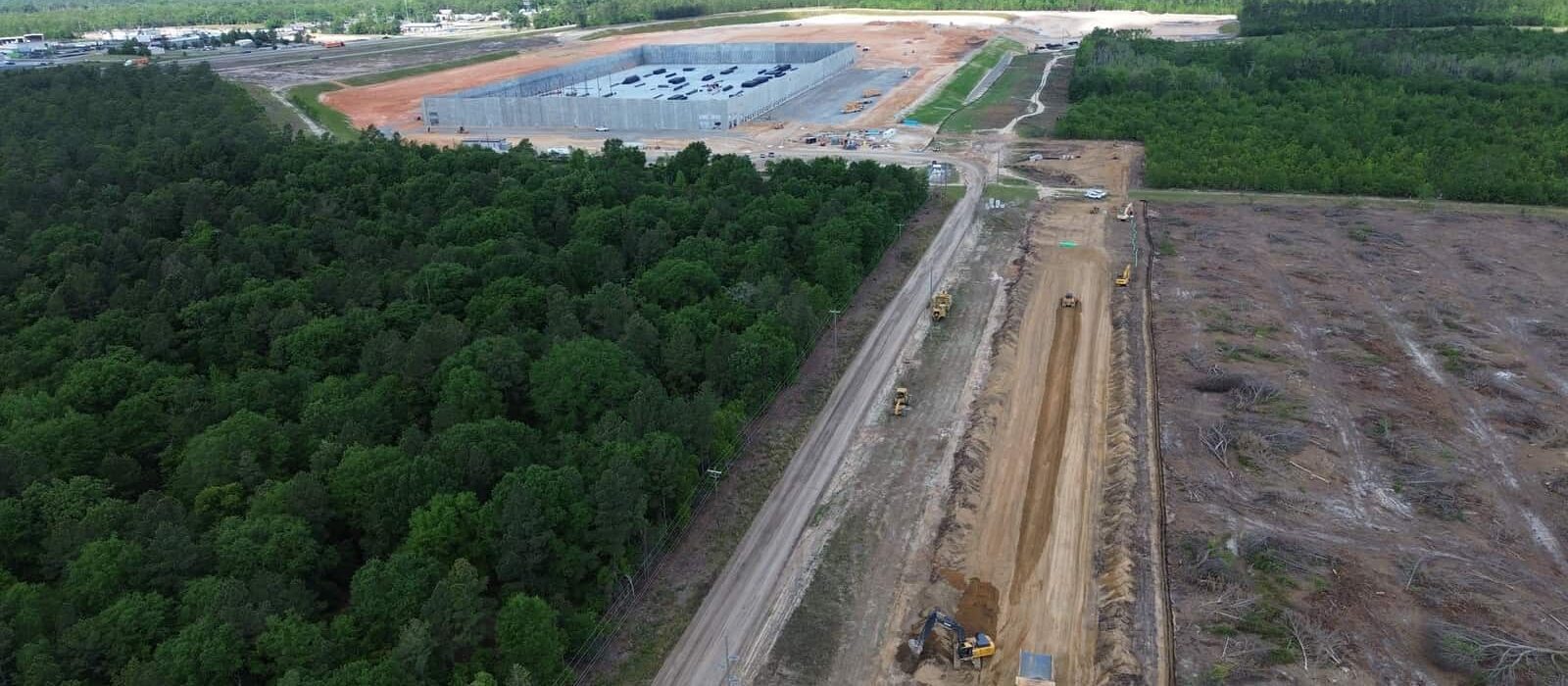 Aerial view of construction near a forested area, showing road building, site prep, and a partially completed industrial building.