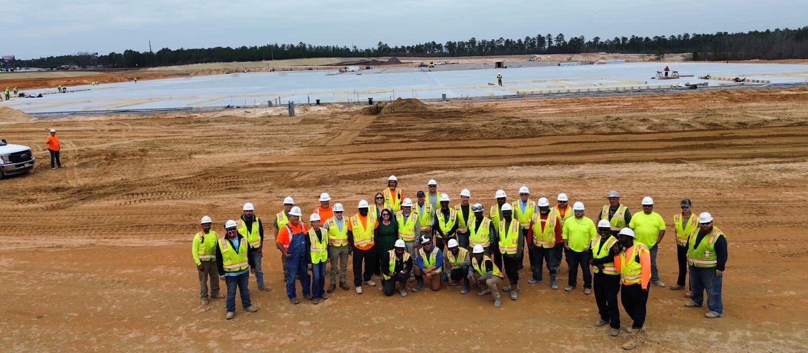 Construction workers and site personnel wearing safety gear posing for a group photo in front of a large slab foundation.