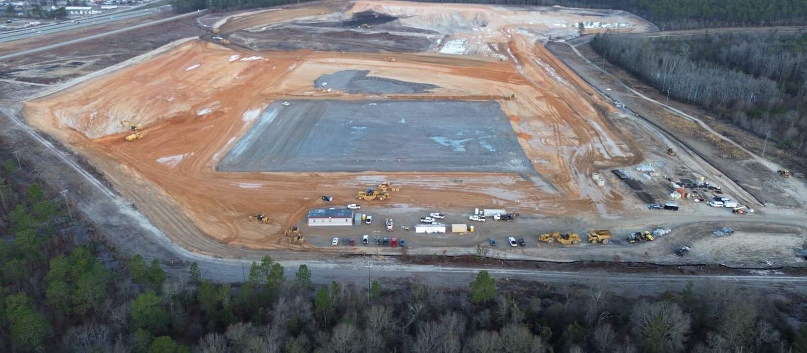 Large-scale construction site with earthwork underway, surrounded by forest, vehicles, and work trailers