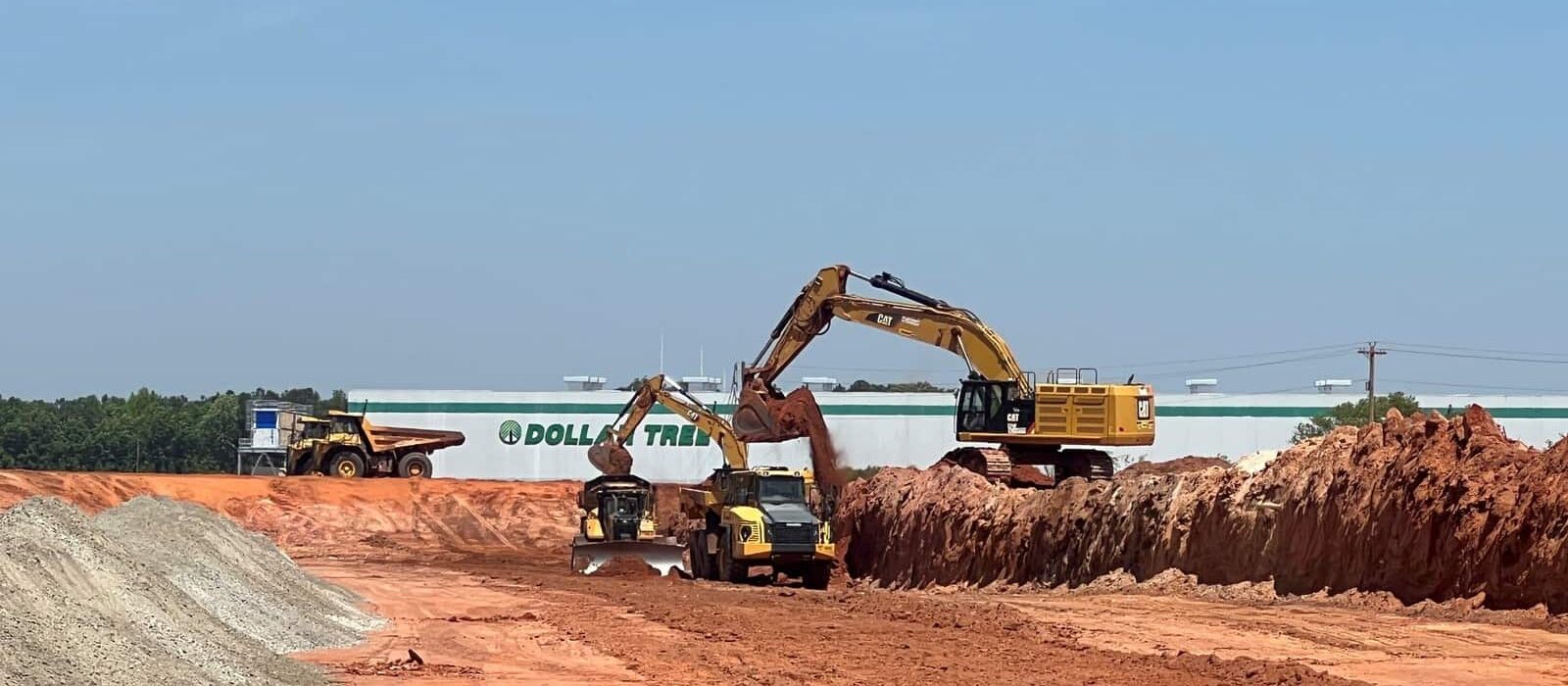 Excavators and dump trucks at a construction site moving red soil, with a Dollar Tree distribution center visible in the background.