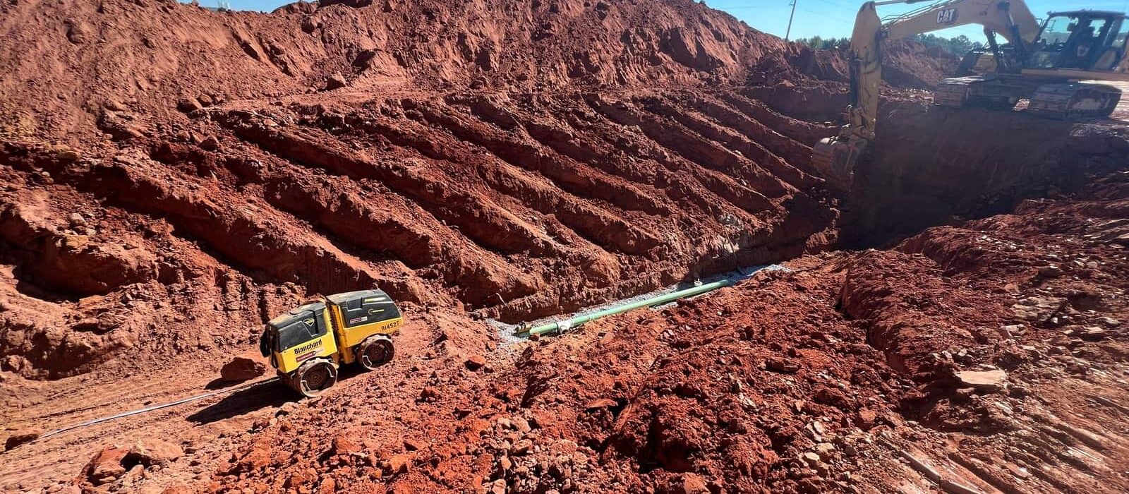 Excavator digging a deep trench with exposed red soil layers and compact roller nearby during utility installation at a construction site.