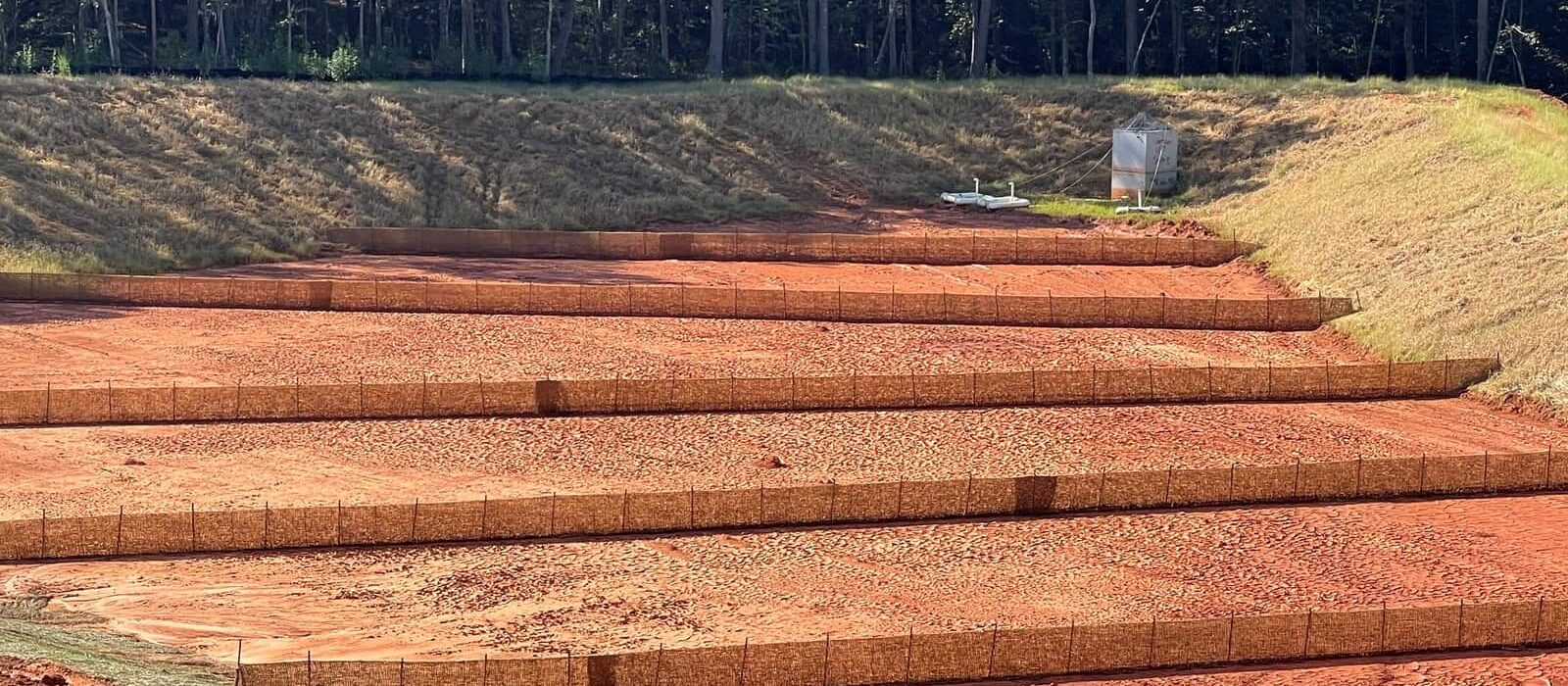 Terraced sediment basin lined with erosion control blankets and silt fences at a construction site near a wooded area.