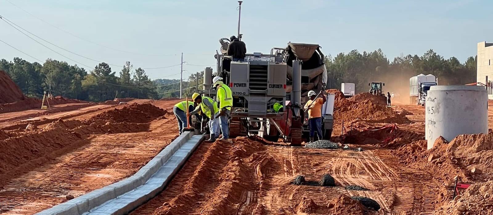 Construction workers operating a curb machine to pour concrete curbing along a red dirt road at an industrial site development.